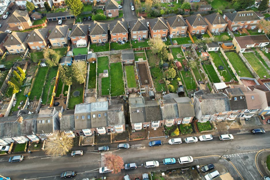 A gardener wearing dark clothing and white gloves is trimming a low hedge with garden shears in a neatly maintained public park area. The scene includes a lawn with slightly damp, green grass and a se